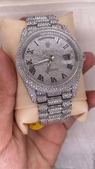Angled view of a replica moissanite watch featuring a stone-covered dial, Roman numerals, and day-date display on a studded strap.