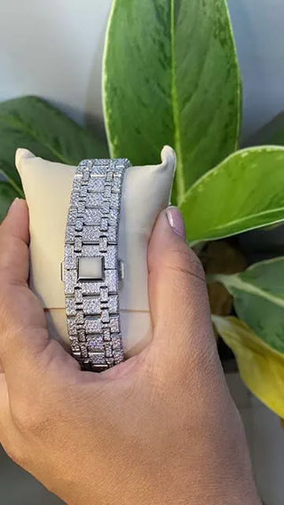 Back view of a men’s VVS moissanite watch, fully iced out with sparkling stones, held in hand with green leaves behind.